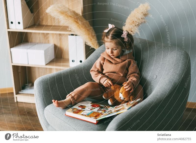 A cute little girl flips through a colorful educational book and sits in a chair in the room. Development, education, childhood sitting toddler happiness home
