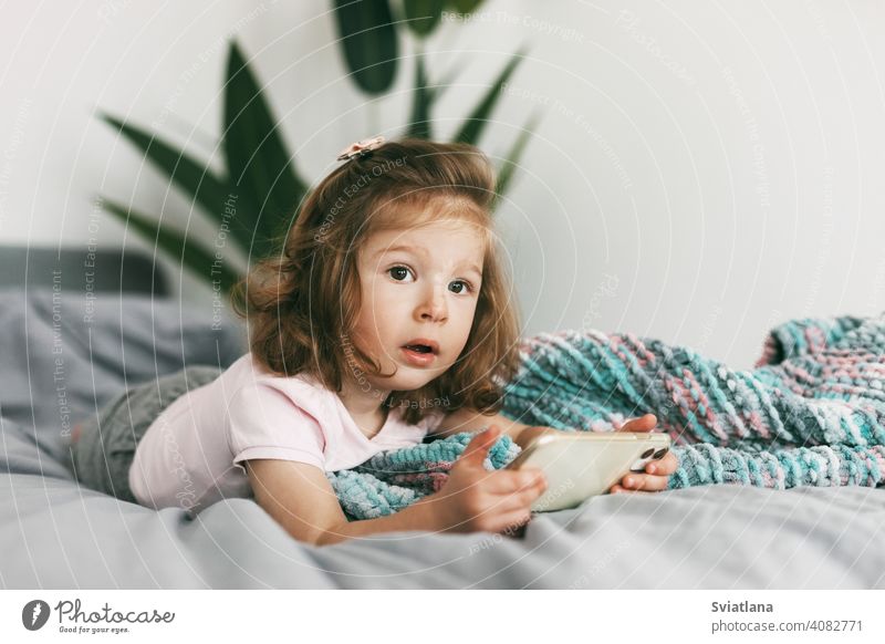 Portrait of a charming little girl who watches cartoons on her phone before going to bed. Childhood, happy time, care smartphone child kid mobile home small