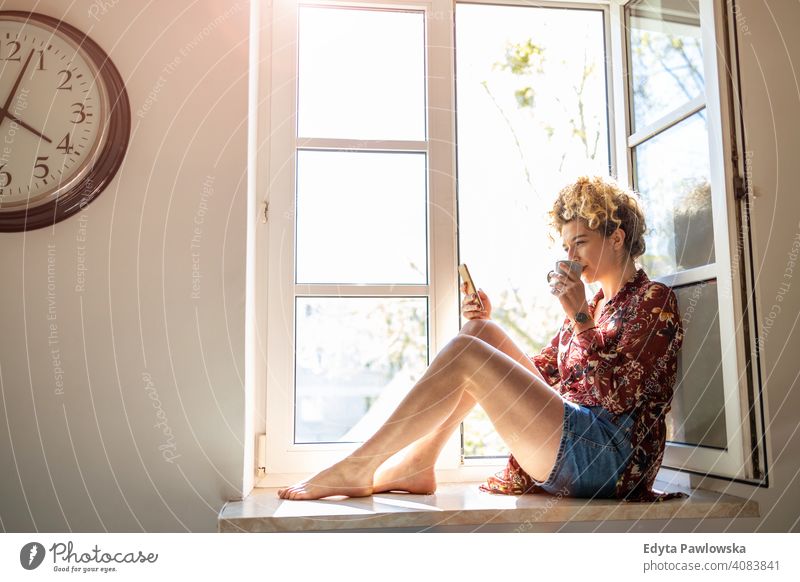 Young woman sitting on window sill and using smartphone happy smiling cheerful apartment leisure bedroom house home alone people caucasian adult indoors person