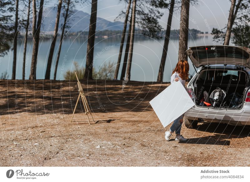 Anonymous female artist with canvas near car countryside lake trunk open easel woman blank modern hobby lifestyle leisure nature sunny daytime casual vehicle
