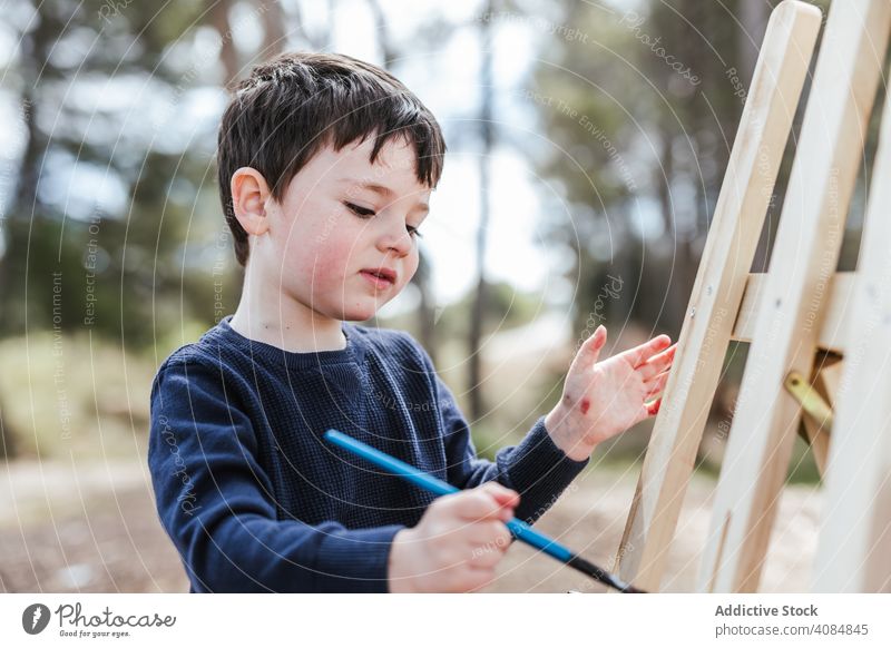 Boy painting on easel in countryside boy canvas art paintbrush child creative kid red leisure lifestyle abstract cute adorable sweet sitting color little nature