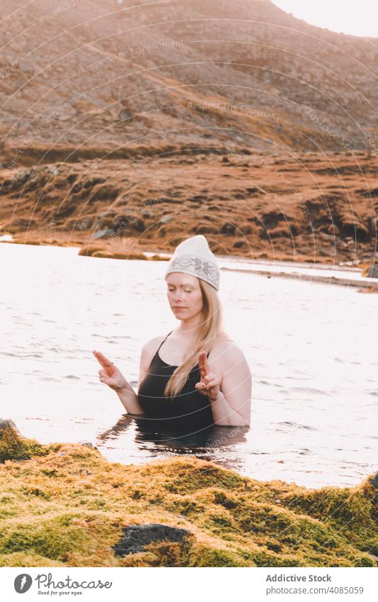 Woman in hat meditating in water near stone shore woman closed eyes swimsuit cold surface coast rock relaxation young nature yoga female hill lifestyle