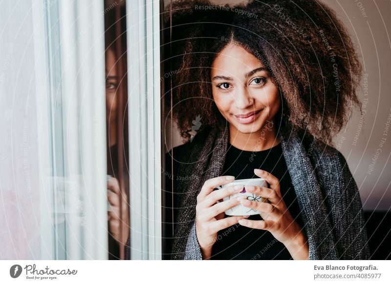 portrait of beautiful afro american young woman by the window holding a cup of coffee. Lifestyle indoors home ethnic mixed race tea autumn winter blanket