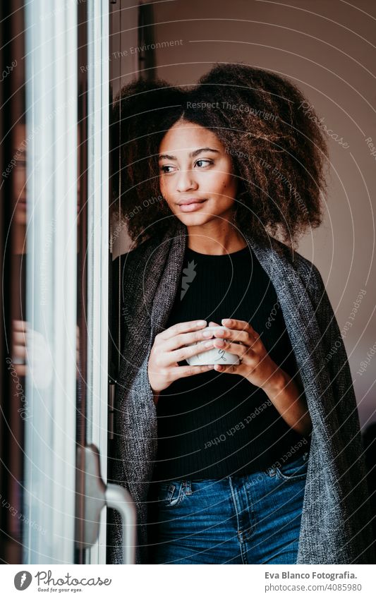 portrait of beautiful afro american young woman by the window holding a cup of coffee. Lifestyle indoors home ethnic mixed race tea autumn winter blanket