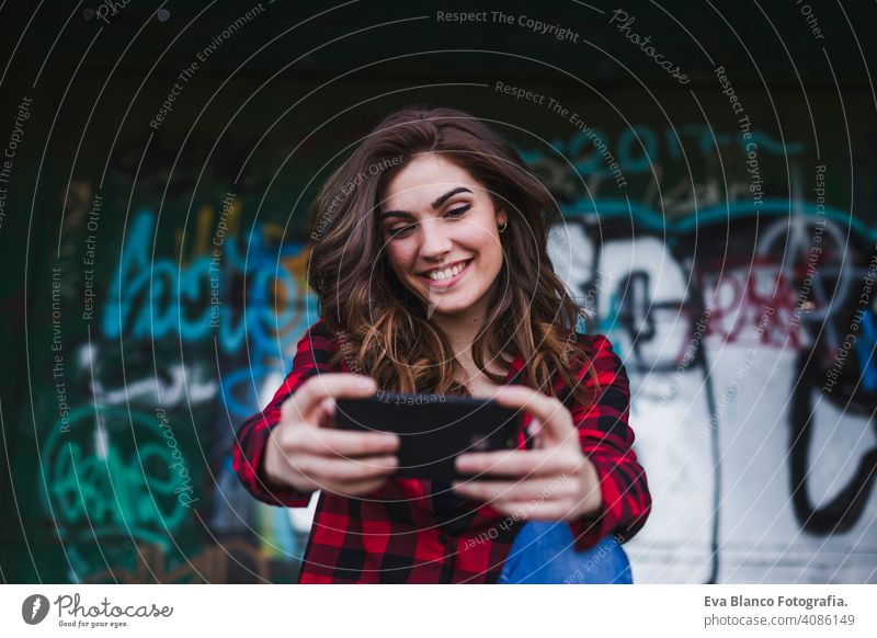 young beautiful woman using mobile phone. She is taking a picture. Urban graffiti background. Outdoors lifestyle.Technology urban hat standing attractive fence