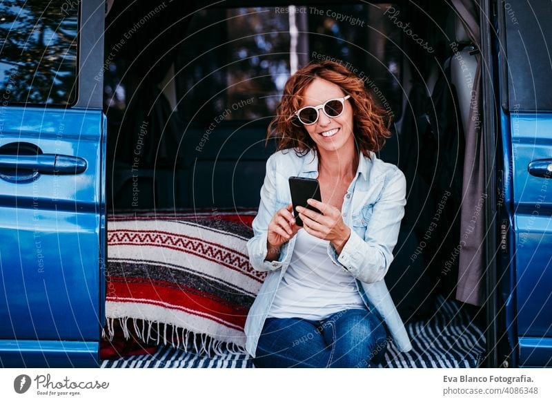happy woman sitting in a blue van and having fun. travel concept. woman using mobile phone laugh car enjoying outdoors resting camp path way sunset wanderlust
