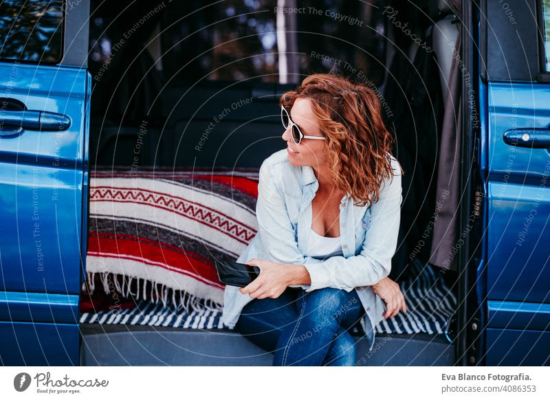 happy woman sitting in a blue van and having fun. travel concept. woman using mobile phone laugh car enjoying outdoors resting camp path way sunset wanderlust