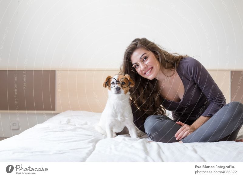 beautiful young woman lying on bed with her cute small dog besides. Home, indoors and lifestyle. They are wearing hipster glasses cozy tired dream mood cuddle