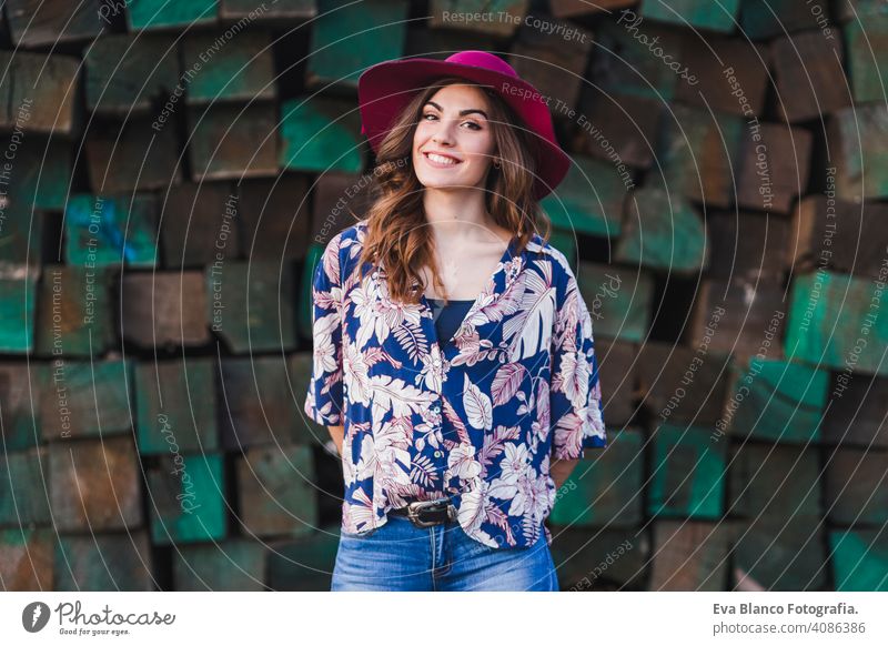 portrait of a young beautiful woman wearing casual clothes and a modern hat, standing over green wood blocks background and smiling. Outdoors lifestyle. youth