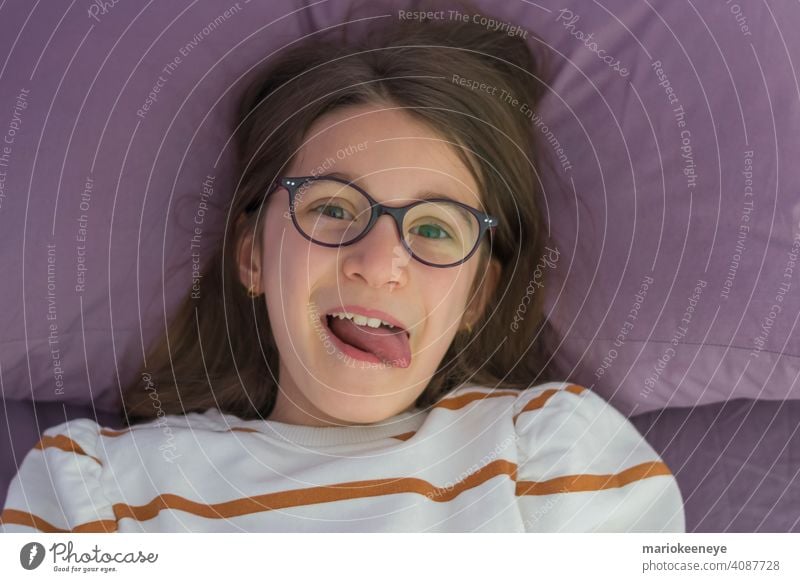 Close-up of little Caucasian girl with glasses lying on bed and sticking out her tongue Vision Horizontal innocence Innocent Laughter Setting jubilant