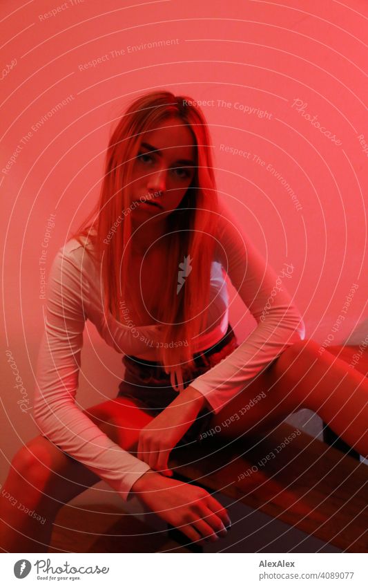 Young woman in hot pants and top sits on a staircase and looks directly into the camera while illuminated with red light Woman young maiden Girl pretty Slim