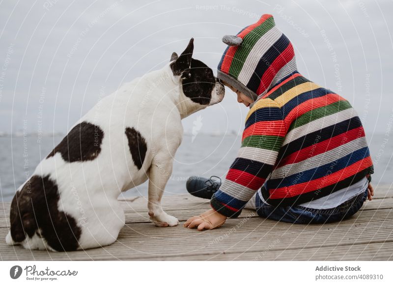 Anonymous kid petting dog on pier patting sea sitting obedient baby french bulldog casual child lifestyle leisure rest relax striped jacket hood puppy loyal