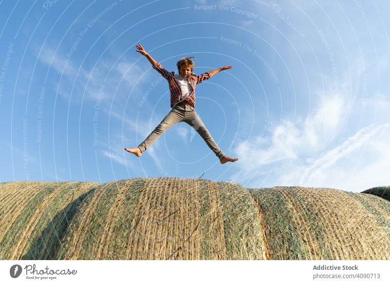 Barefoot boy jumping on haystacks farm sky clouds sunny daytime nature lifestyle leisure teen kid child barefoot dried grass straw rolls countryside rustic