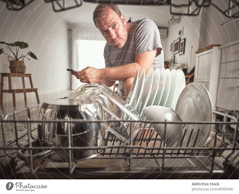 Angry adult man with smartphone while repairing dishwasher in kitchen father fix tools angry family help equipment dishware broken damaged defective teach skill