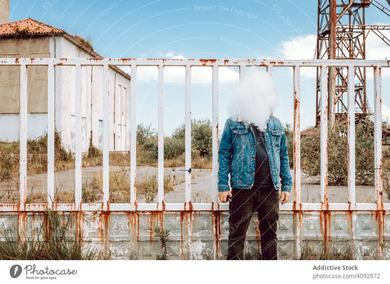 Anonymous vaper standing near rusty barrier man cloud fence derelict building industrial exhale habit male hipster e-cigarette vapor cool smoking nicotine