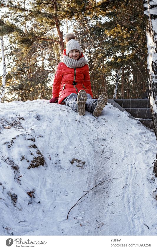 Young smiling cute girl in a bright sport suit is preparing to sled down the hill. Happy childhood, winter outdoor activity. Vertical shot young sledding
