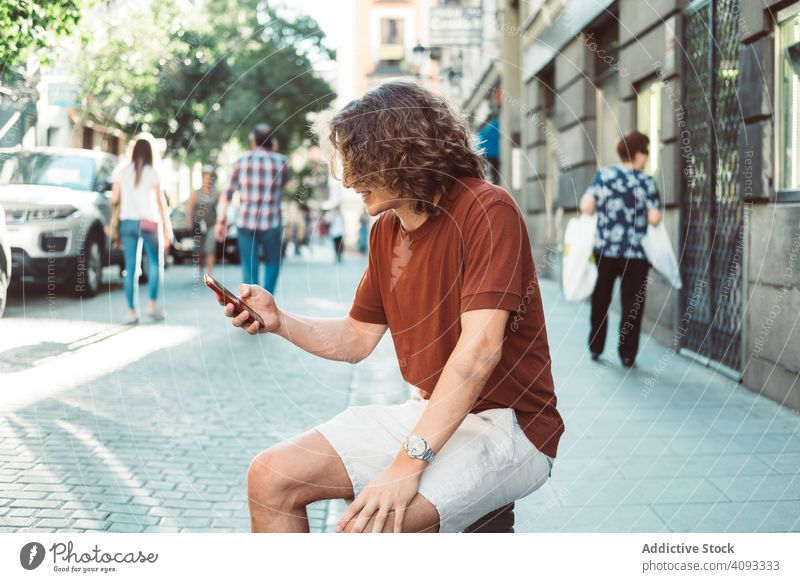Amused man with longboard talking on phone while resting on street amused casual laugh gesture cheerful using speak smartphone sunny baluster pavement sit