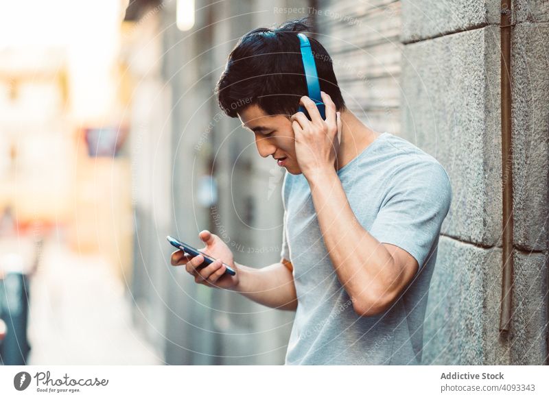 Content Asian man in headphones listening music on smartphone on street city using streaming content cool joyful modern casual stand sunny town young adult