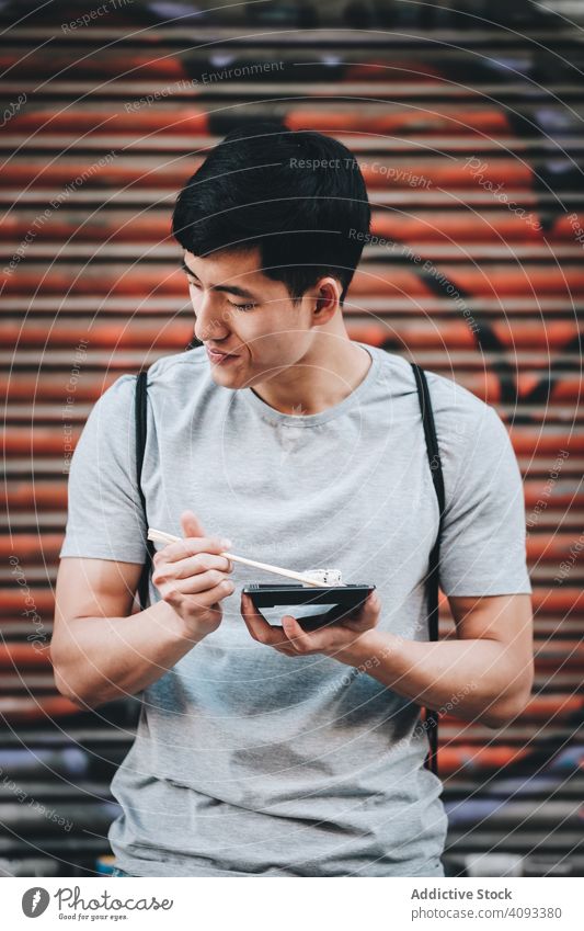 Asian man eating takeaway food with chopsticks on city street street food hungry asian food urban sunny food truck enjoying ethnic fast food young adult meal