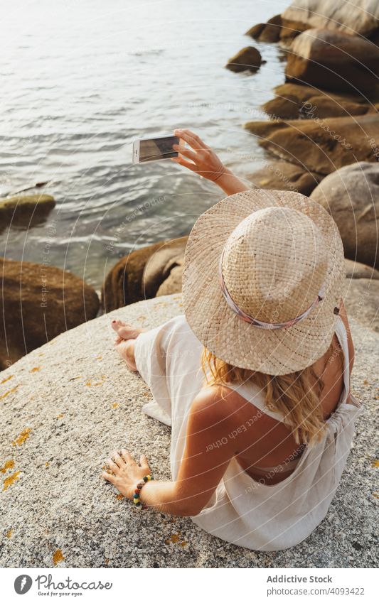 Contemporary woman with phone chilling on seashore smartphone coast alone enjoy escape travel seaside solitude remote wireless straw hat gadget trip vacation