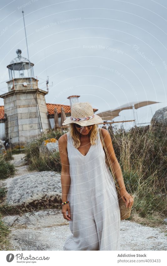 Woman in hat standing against beacon on seashore woman lighthouse ocean stone coast rock architecture walk building grass travel trip gloomy vacation cloud