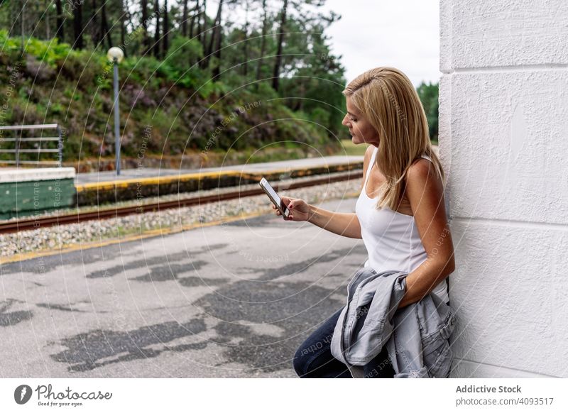 Contented woman staying on railway station with smartphone using railroad watching browsing platform gadget device surfing travel waiting tourist journey online