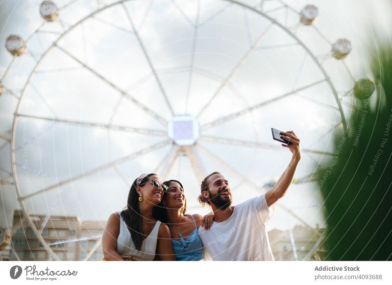 Carefree friends taking selfie on smartphone while standing beside carousel at funfair carefree cheerful smile having fun tanned photo hug attraction carnival