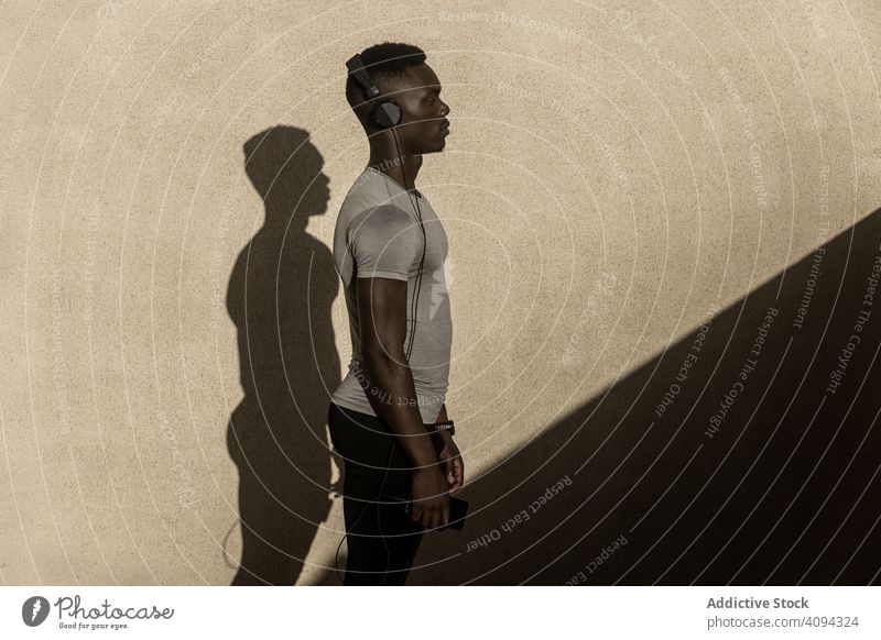 Ethnic sportsman in headphones standing against gray wall silhouette shadow posture confident ethnic athletic muscle workout sportswear stamina fit solitude