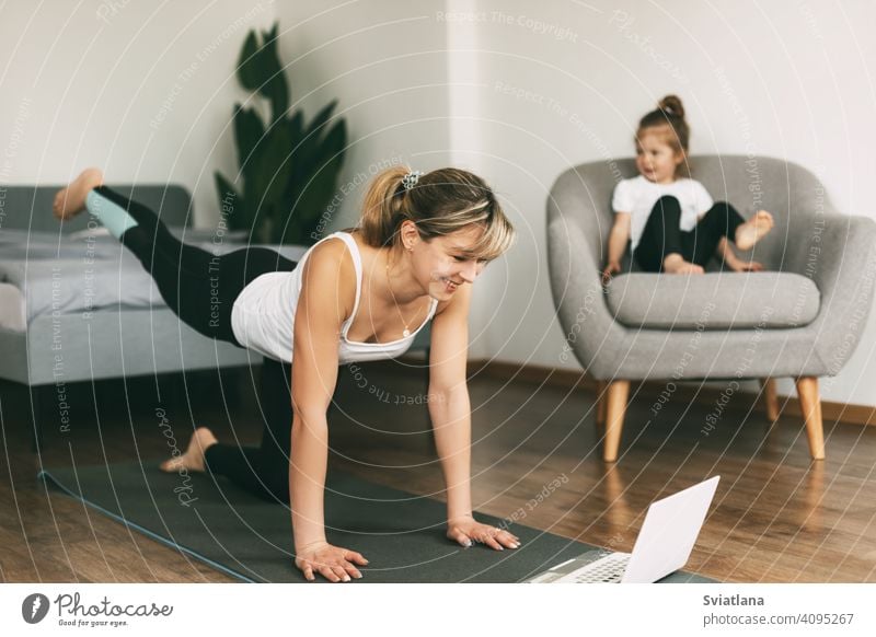 A young mom is doing sports at home and watching a video fitness lesson on a laptop, and her little baby is sitting on a chair. Home training, fitness, sports