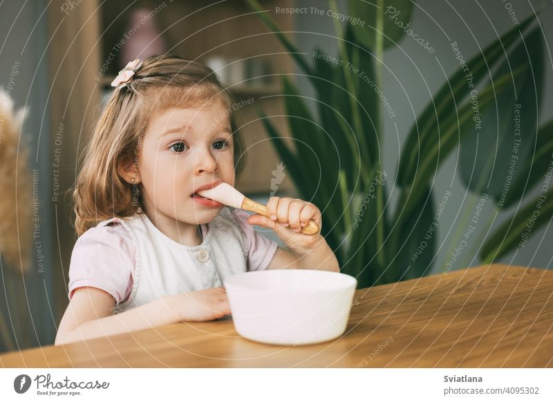 A little girl eats porridge from a white bowl, a girl has breakfast sitting at the table. Healthy breakfast, healthy food child eating plate baby cute childhood