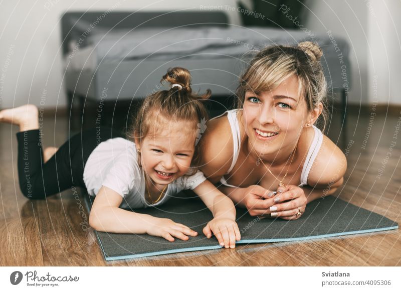 A beautiful young woman and an adorable little girl look at the camera and smile while lying on a yoga mat in the bedroom fitness smiling body training