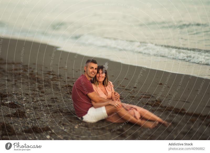 Delighted couple sitting on beach resort love hug date sea smile happy vacation man woman adult honeymoon summer shore coast relationship water embrace ocean