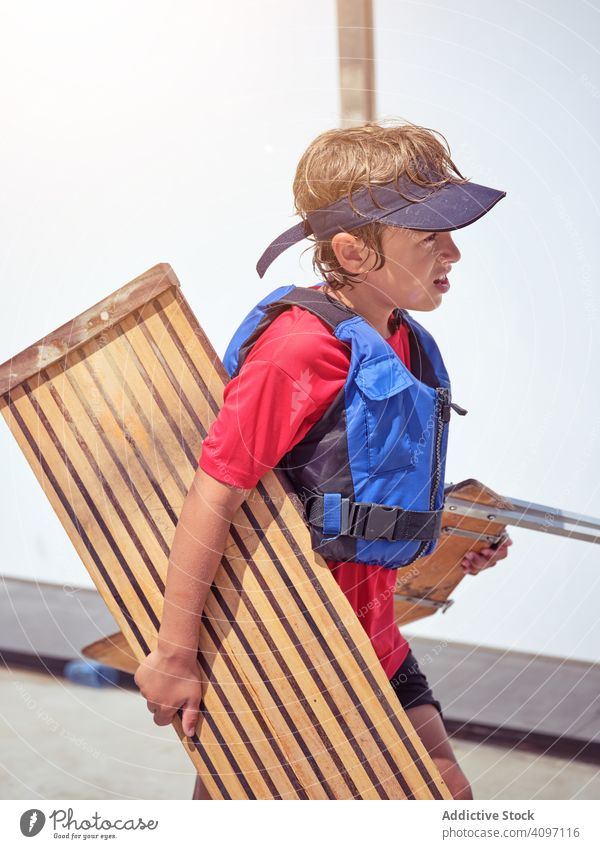 Skilled boy in blue life jacket walking with wooden stand swimming yacht kid cruising water shore sport wind ocean holiday tense childhood hobby fun wet