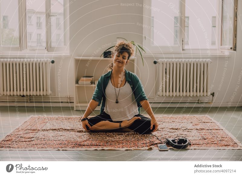 Peaceful woman meditating in lotus position on floor at home peaceful relaxed smile casual carpet apartment modern smartphone headphones young adult happy yoga