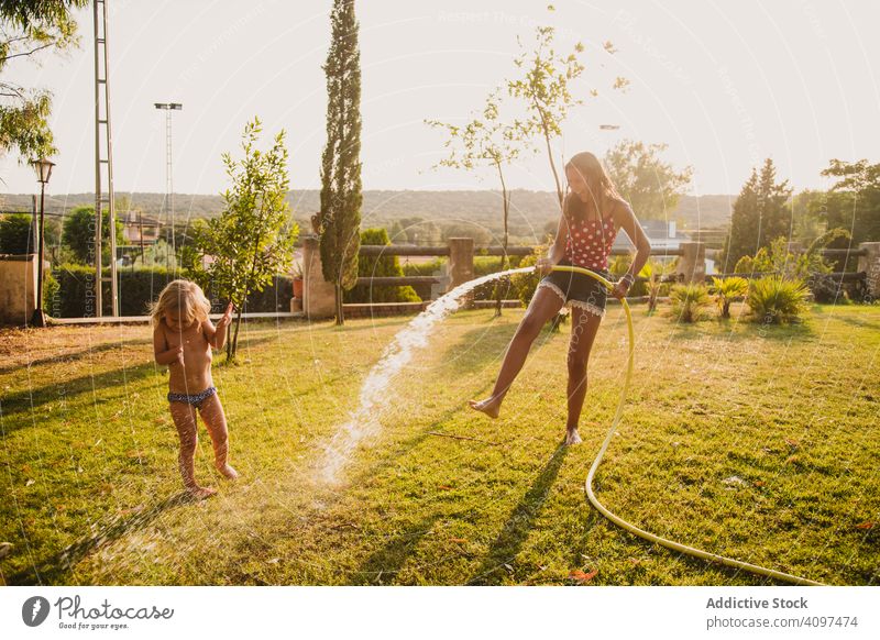 Teen girl spraying little sister in garden fun water lawn hose sunny daytime yard together teen kid child happy cheerful play vacation weekend lifestyle leisure