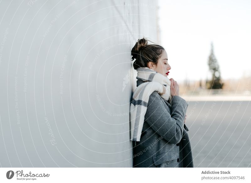 Young woman in coat and scarf on windy day stylish street closed eyes wall city building female urban fashion cool young model outfit warm weather exterior lady