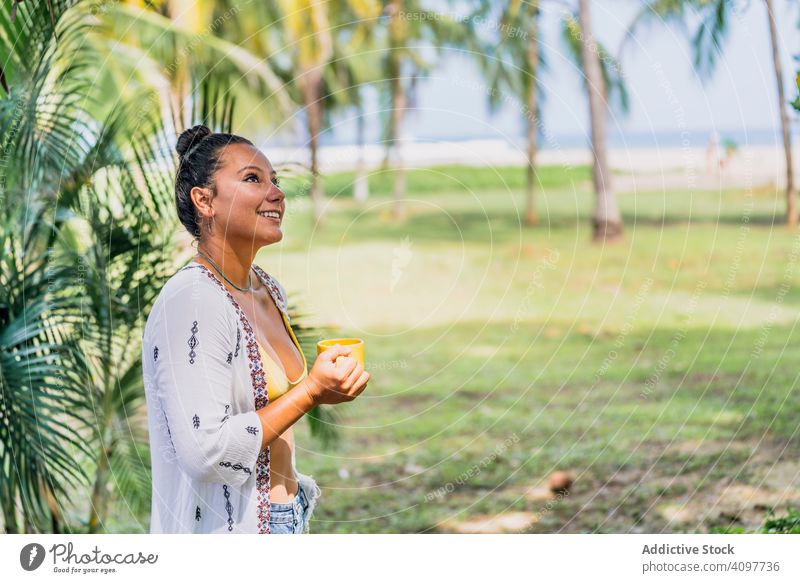 Relaxed woman enjoying hot drink and standing on exotic lawn relaxed seaside palm tree sunny peaceful coffee mug grass tropical rest costa rica young tanned