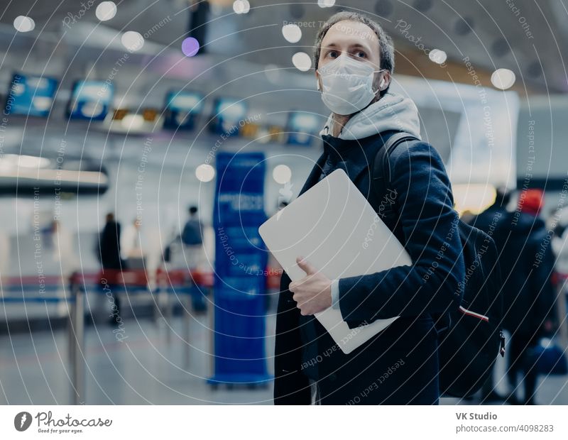 Man traveler wears protective disposable medical mask in airport, returns from abroad where coronavirus spreading, carries backpack, takes care of health, protects from virus, has postponed flight