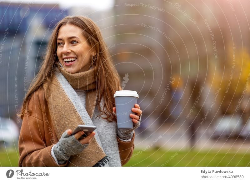 Smiling young woman with smart phone and coffee cup outdoors at urban setting autumn fall girl female beautiful weather coat fashion pretty attractive adult