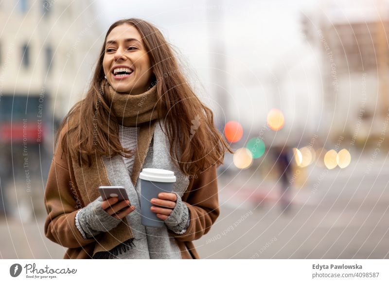 Smiling young woman with smart phone and coffee cup outdoors at urban setting autumn fall girl female beautiful weather coat fashion pretty attractive adult