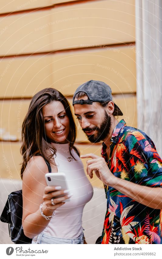Trendy ethnic man and woman sharing mobile phone checking photos while standing on street friend share urban stroll trendy together couple smartphone modern