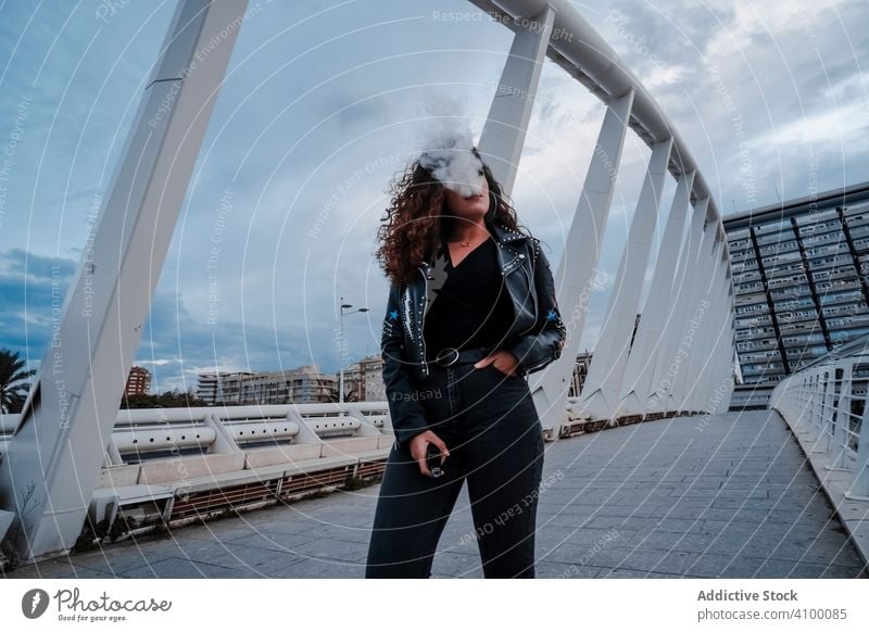 Stylish woman in black jacket and jeans smoking on bridge urban leather rock city style stripes street brunette fashion outfit young female underground clothes