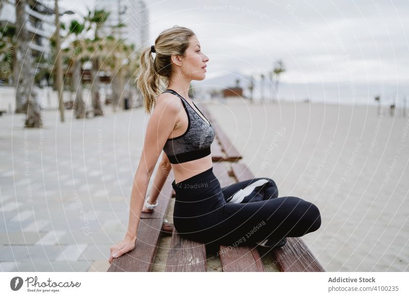 Sportswoman resting in a bench at sandy beach stretching exercising arm hands sitting cross legs fitness healthy workout athlete sportswear female young
