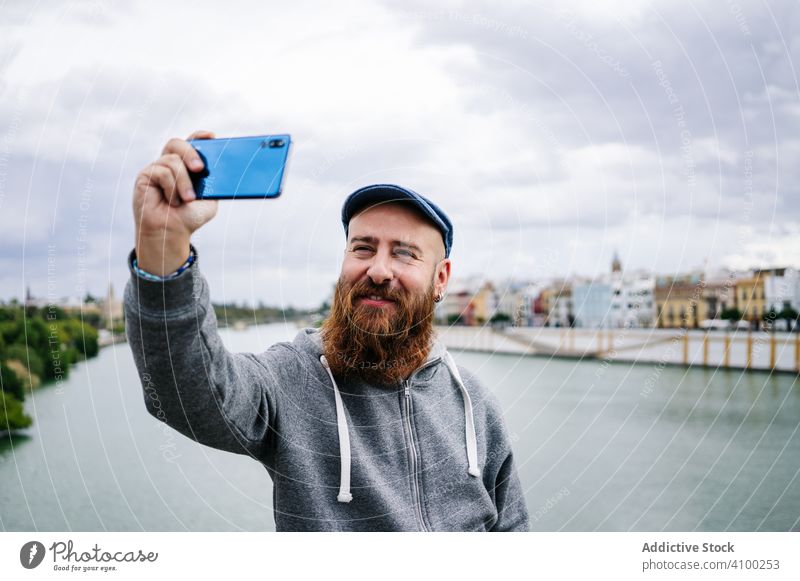 Satisfied male tourist taking selfie on smartphone at quay making photo mobile using browsing peace sign holding device gadget casual bearded river bay bridge