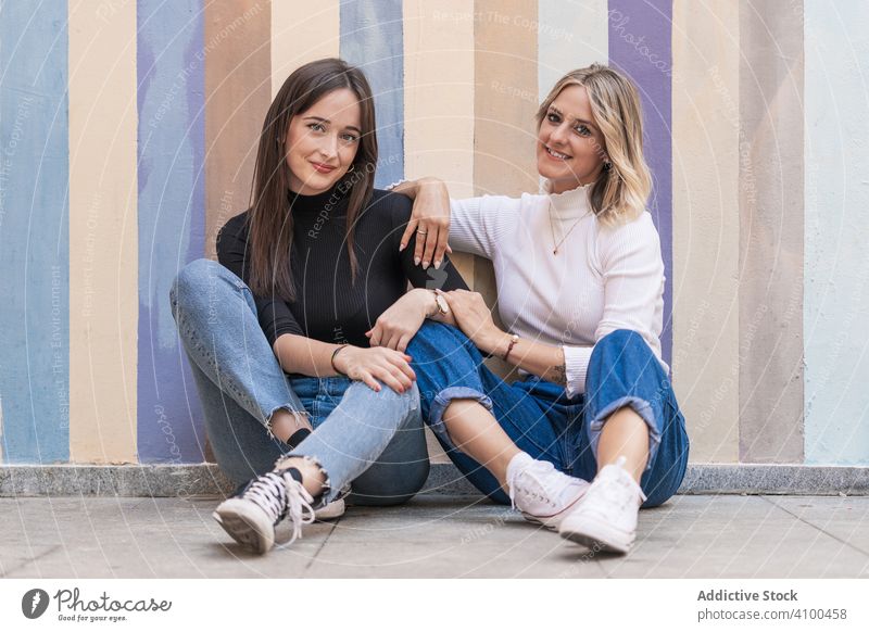 Happy girlfriends sitting against painted urban wall together women friendship touch trendy cheerful positive stylish relationship elegant street cool colored