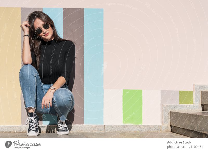 Trendy female sitting at street against colored wall trendy woman charming cheerful elegant stylish glamour positive carefree gorgeous urban appearance content