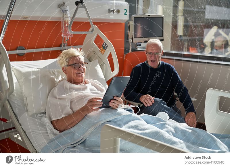 Senior couple using devices in hospital ward patient visitor tablet smartphone senior medical care man woman married bed clinic room healthcare gadget