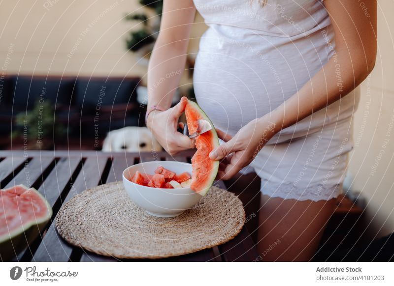 Faceless pregnant woman cutting watermelon to bowl summer expectancy table terrace watch motherhood morning rest female fresh juicy dessert slice lady nutrition