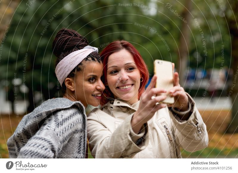Cheerful multiracial girlfriends taking selfie on smartphone in park couple smiling relationship together cheerful vacation gadget device fashion autumn women