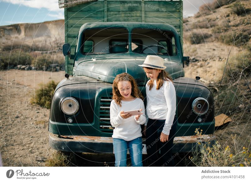 Cheerful girls watching smartphone near abandoned truck children car focused rest laugh smile cheerful using jumper casual share nature relax desert